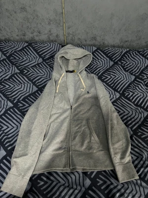 Grå hoodie med dragkedja från Polo - Snygg grå hoodie från Polo Ralph Lauren med vit snörning i huvan och liten blå logga på bröstet. Tröjan har dragkedja framtill, två fickor och långa ärmar. Perfekt för chill dagar och enkel att matcha med jeans eller joggers.
