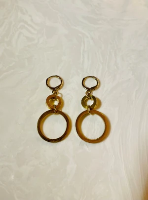 Guldiga cirkelörhängen i stål - Säljer ett par trendiga örhängen i guldtonad metall med två cirklar i olika storlekar som hänger i varandra. De har en enkel men cool design som verkligen sticker ut och passar perfekt om du vill addera lite extra stil till din look.