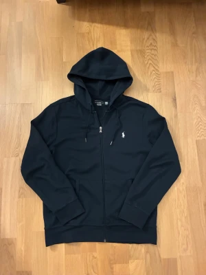 Svart hoodie från Polo Ralph Lauren - Svart hoodie från Polo Ralph Lauren i storlek M. Tröjan har huva med snörning, dragkedja framtill och två fickor. Klassisk broderad logga i vitt på bröstet. Tillverkad i mjuk bomull, perfekt för chill dagar och sköna outfits.