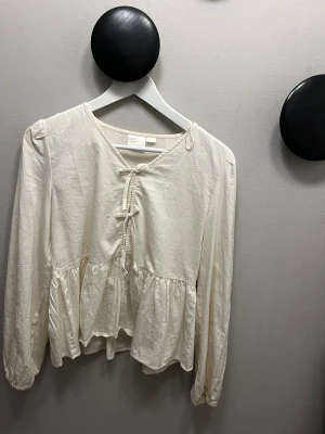 Krämvit blus med rosetter och volang - Säljer en krämvit blus från gina Tricot med långa ärmar och två rosetterna framtill. Blusen har volang nedtill och är gjord i mjuk bomull. Perfekt för dig som gillar romantiska detaljer och en avslappnad vibe.