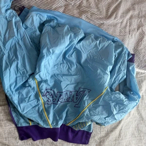 Nike Lakers vindjacka blå/lila M - Säljer en sjukt snygg Lakers vindjacka från Nike i blått med lila detaljer och gula inslag. Jackan har dragkedja, meshfoder och NBA-logga. Perfekt för dig som älskar basket och vill sticka ut med stilren sportig look.