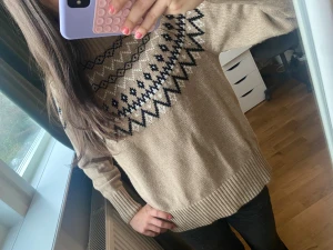 Beige stickad tröja med mönster - Snygg beige stickad tröja med svart och vitt geometriskt mönster över axlar och bröst. Tröjan har ribbad hals, muddar och nederkant samt långärmad passform. Perfekt för dig som gillar mysiga och trendiga vinterlooks.