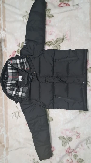 Svart dunjacka/väst från Burberry, M - Svart dunjacka från Burberry med klassiskt rutigt foder i grått, vitt och rött. Jackan har huva, dragkedja och tryckknappar framtill samt två fickor med dragkedja. Perfekt för kalla dagar och riktigt snygg streetstil. Man kan ta av armarna och göra den till en väst, Helt my e etikettet på oanvänd 