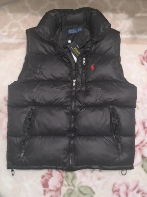 Svart dunväst från Polo Ralph Lauren - Svart dunväst från Polo Ralph Lauren med hög krage, tre dragkedjefickor och klassisk röd logga broderad på bröstet. Västen är quiltad och har en clean, sportig look med glänsande yta. Perfekt för lager-på-lager och streetstyle.helt ny med etikett på