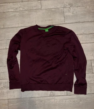 Hugo Boss - Snygg vinröd sweatshirt från Hugo Boss med diskret logga på bröstet. Tröjan har ribbade detaljer på axel och sidor samt långa ärmar. Perfekt för dig som gillar stilrena och moderna plagg med lite edge. (Finns ett litet hål på baksidan som knappt syns) Nypris:899:-