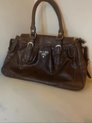 Vintage Prada väska - Snygg brun handväska från Prada i äkta skinn med klassisk form och silverfärgade detaljer. Väskan har dubbla handtag, justerbar axelrem och praktisk dragkedja upptill. Insidan är rymlig med innerfack och logotyp. Perfekt för dig som vill ha en stilren och lyxig väska. Pris går att diskutera och vid snabb affär kan jag gå ner i pris☺️