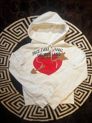 Vit hoodie Helmut Lang  - Vit hoodie från Helmut Lang med stor röd hjärtdetalj och pil på bröstet samt svart text. Tröjan har huva med snörning och klassisk känguruficka❤️| Skriv vid funderingar!          Priset kan självklart diskuteras!