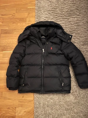 Svart pufferjacka från Polo Ralph Lauren - Svart pufferjacka från Polo Ralph Lauren med huva och två dragkedjeförsedda fickor. Jackan har en röd Polo-logga på bröstet och grov dragkedja framtill. Perfekt för kalla dagar och har en schysst, puffig look. Priset är inte fast.