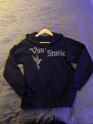 Svart hoodie Von Studio med dragkedja - Svart hoodie från Von Studio med glittrande text och kolibri på framsidan. Tröjan har huva, långa ärmar och praktisk dragkedja framtill. Perfekt för dig som gillar streetwear och vill sticka ut med snygga detaljer. Passar dig som är 165