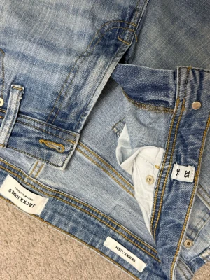 Jack & Jones ljusblå skinny jeans - Säljer ett par ljusblå skinny jeans från Jack & Jones med klassisk femficksdesign och slitna detaljer. Jeansen har gul kontrastsöm och är tillverkade i stretchigt denim för en tajt passform. Perfekt för dig som gillar en smal siluett.