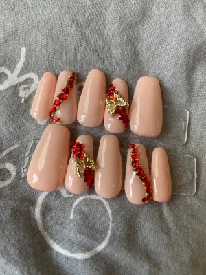 Nude lösnaglar med röda stenar - Snygga lösnaglar i nude färg med glansig finish. Dekorerade med röda och guldiga stenar samt små guldfärgade fjärilar. Naglarna är långa och har en rak, kistformad design. Perfekt för dig som vill ha en extra glam look.