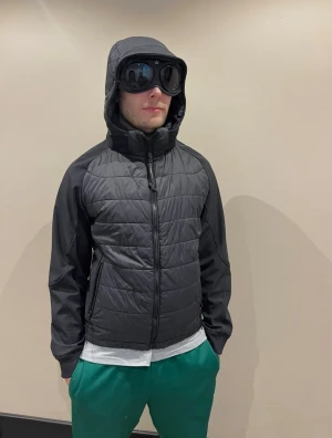 Svart pufferjacka från C.P. Company - Svart pufferjacka från C.P. Company med huva och hel dragkedja framtill. Jackan har quiltad front och rygg, släta ärmar och två sidofickor med dragkedja. Perfekt för kyliga dagar och har en clean, sportig look. Modellen är 183cm lång. Storleken är L/52.