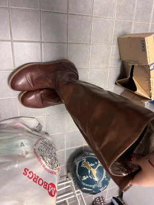 Bruna höga stövlar i skinn - Snygga bruna stövlar i äkta skinn med rund tå och låg klack. De har dragkedja på insidan och en klassisk, stilren look som passar perfekt till jeans eller kjol. Sulan är mönstrad för extra grepp och stövlarna går upp till knäet.