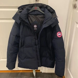Mörkblå dunjacka från Canada Goose - Säljer en mörkblå dunjacka från Canada Goose med huva och klassisk logga på ärmen. Jackan har dragkedja och knappar framtill, samt en clean och puffig design som håller dig varm under kalla dagar. Perfekt för dig som vill ha en snygg och funktionell vinterjacka.