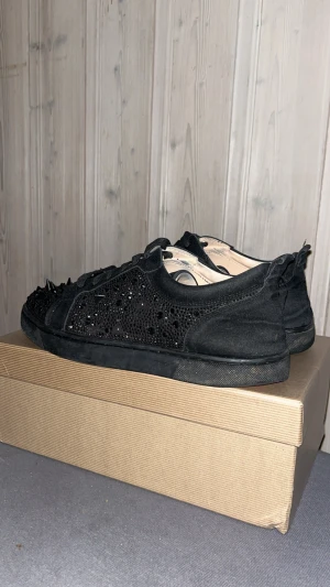 Christian Louboutin svarta sneakers med nitar - Svarta sneakers från Christian Louboutin med ikonisk röd sula, dekorerade med svarta nitar och glittrande detaljer på ovansidan. Skorna har snörning och är tillverkade i mocka och syntetmaterial. Snygg, edgy look med rund tå och platt sula.