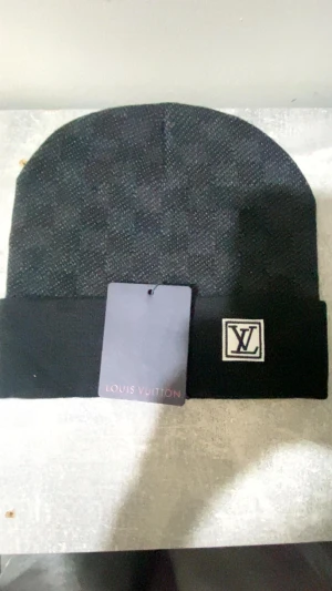 Svart Louis Vuitton mössa med logga - Snygg svart mössa från Louis Vuitton med klassisk LV-logga framtill på uppvikt kant. Mössan har en stilren och enkel design, perfekt för dig som gillar exklusiva märken. Materialet är stickat och känns mjukt mot huvudet. Taggen sitter kvar för extra lyxig känsla.