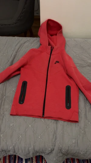 Röd hoodie från Nike med dragkedja - Nike hoodie i rött med svart logga på bröstet. Tröjan har hel dragkedja framtill, två svarta fickor med dragkedja och en stor huva. Materialet är mjukt och känns som bomull eller polyester. Perfekt för chill eller träning.