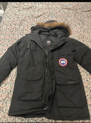 Svart dunjacka från Canada Goose - Svart dunjacka från Canada Goose med huva och pälsdetalj, klassisk logga på bröstet och stora fickor framtill. Jackan har dragkedja och är långärmad, perfekt för kalla vinterdagar. Snygg och funktionell design med broderad patch och märkning på ryggen.