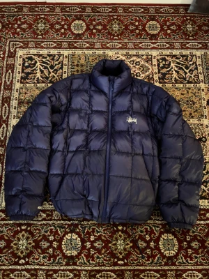 Stussy midweight puffer jacka - En nästan helt ny stussy puffer jacka, knappast använd men den har ett jättelitet hål vid ryggen, men det är inget man tänker på! Den är mörkblå/lila. Skriv om ni har några frågor!☺️