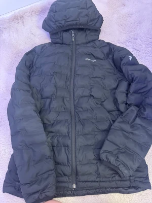 Svart pufferjacka från Peak Performance - Svart pufferjacka från Peak Performance med huva och hel dragkedja framtill. Jackan har quiltad design och logga på bröstet. Perfekt för kalla dagar med sin fluffiga och isolerande känsla. Snygg och enkel stil som funkar till det mesta.