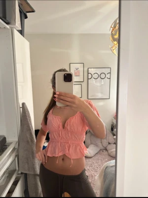 Korall blus - Supersöt korall blus.Toppen har korta puffärmar som kan bäras både på axeln och offshoulder. Den är croppad med volangkant nertill och har en feminin v-ringning. Perfekt för dig som gillar romantisk och trendig stil.