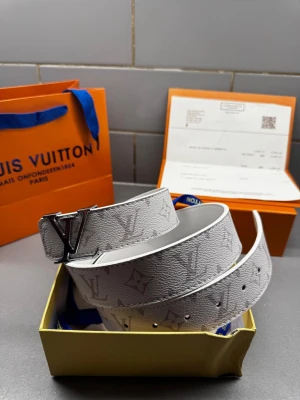 Louis Vuitton vitt skärp med monogram - Säljer ett vitt Louis Vuitton-bälte i skinn med klassiskt LV-monogram i ljusgrått. Bältet har en bred design och en stor, silverfärgad LV-logga som spänne. Snygg struktur i lädret och exklusiv känsla, perfekt för att lyfta vilken outfit som helst.