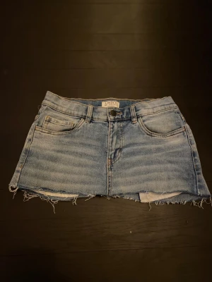 Blå jeanskjol från Lindex - Snygg blå jeanskjol från Lindex med klassisk femficksdesign och rå kant nedtill för en cool, avslappnad vibe. Kjolen har knapp och dragkedja framtill samt ljusa tvättade detaljer som ger en fräsch look.