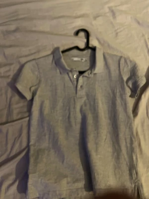 Ljusgrå pikétröja från 157 - Säljer en stilren ljusgrå pikétröja från 157 med klassisk krage och knappar framtill. Tröjan är kortärmad och tillverkad i mjuk bomull, perfekt för en clean och avslappnad look.