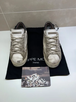 Philippe Model vita sneakers i skinn - Snygga vita sneakers från Philippe Model Paris med beige och grå detaljer. Skorna har klassisk låg profil, rund tå och snörning. Tillverkade i skinn och mocka med kontrasterande hälparti i mörkgrå mocka. Perfekta för dig som gillar en stilren men avslappnad look.