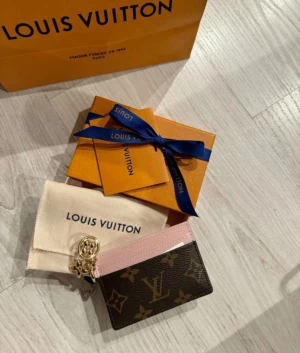 Louis Vuitton plånbok med rosa detalj - Snygg plånbok från Louis Vuitton med klassiskt monogrammönster i brunt och beige, samt en ljusrosa kant upptill. Plånboken har guldfärgad charm och flera fack för kort. Materialet är skinn och textil, perfekt för dig som gillar lyxiga accessoarer.