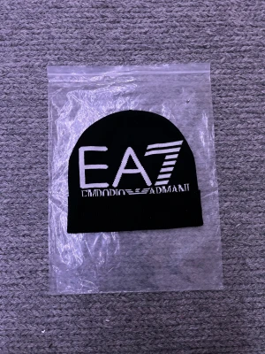 Svart EA7 Emporio Armani mössa - Svart mössa från EA7 Emporio Armani med stor vit logga framtill. Klassisk rundad form med uppvikt kant och ribbad nederdel. Materialet är stickad bomull, vilket gör den mjuk och skön. Perfekt för dig som vill ha en stilren och trendig accessoar.