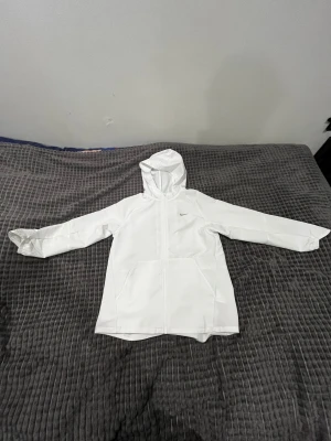 Vit regnjacka från Nike med huva/ Vindjacka  - Nike regnjacka i vitt med minimalistisk design och diskret logga på bröstet. Jackan har huva, dragkedja framtill och två stora fickor. Tillverkad i lätt och vattenavvisande syntetmaterial, perfekt för blöta dagar. Lång ärm och enkel passform. Armarna sitter bra i slutet är dom formade lite de modellen, Använt 1 gång ute men är lite smutsig 