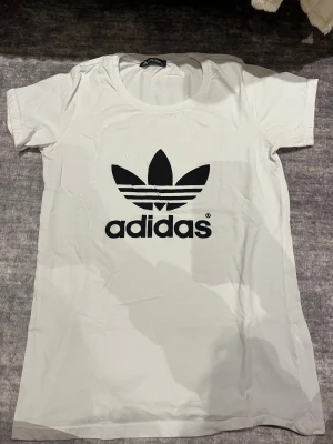 Vit Adidas t-shirt med svart logga - Vit t-shirt från Adidas med den klassiska svarta trefoil-loggan tryckt framtill. T-shirten har rund halsringning och korta ärmar. Tillverkad i mjuk bomull för en skön känsla och enkel stil.