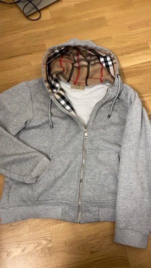 Grå teddyjacka från Burberry XL - Grå teddyjacka från Burberry i storlek XL med dragkedja och huva. Insidan av huvan har det klassiska Burberry-rutiga mönstret i beige, svart och rött. Jackan är fodrad med mjuk teddy och har snörning i huvan samt ribbade muddar. De står att de storlek xl med de passar som L