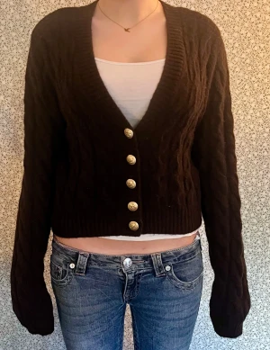 Mörkbrun croppad kofta med guldknappar - Snygg mörkbrun kofta med croppad passform och grovstickat mönster. Koftan har djup v-ringning och fyra dekorativa guldknappar framtill. Långa ärmar och bred ribbad kant vid hals och nederdel. Perfekt för lager-på-lager under kyliga dagar. Aldrig använd.
