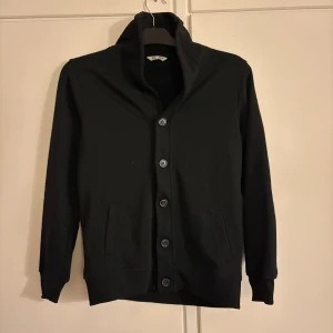 Svart Cardigan - Svart kofta från Cubus i storlek 158/164, sitter aningen större enligt mig. Den har en hög krage, fem knappar framtill och två fickor på sidorna. Skick: 8/10, Nypris: 599kr, Mitt pris: 199kr