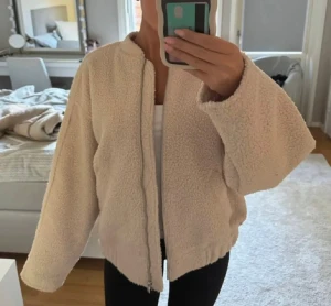 Beige teddyjacka med dragkedja - Mysig beige teddyjacka med dragkedja framtill och avslappnad passform. Jackan har lång ärm och är tillverkad i ett fluffigt fleece-liknande material som ger en cool och trendig look. Perfekt att slänga på sig över en hoodie eller crop top.