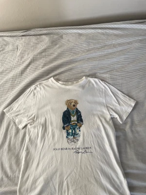 Vit Polo Bear t-shirt från Ralph Lauren - Säljer en vit t-shirt från Ralph Lauren med Polo Bear-tryck på framsidan. T-shirten har rund hals och korta ärmar. Materialet är mjuk bomull och motivet visar en nalle i cool outfit. Perfekt för dig som gillar klassisk streetstyle med en twist.