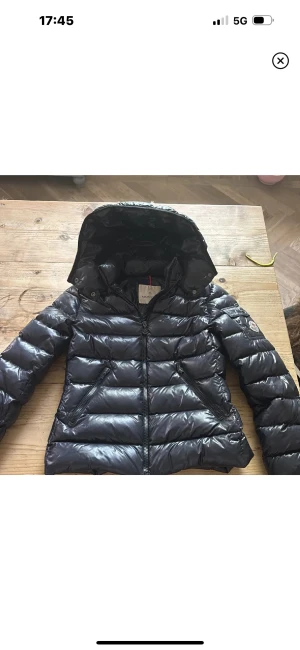 Mörkblå Moncler pufferjacka med huva - Snygg mörkblå pufferjacka från Moncler med glansig finish och klassisk logga på ärmen. Trasig dragkedja längst upp så brukar inte märka av det🫶🏼 Passar XS/S men är storlek 12 i barnstorlek❤️❤️ Ankserien borta inuti men har qr kod för äkthetsbevis🫶🏼