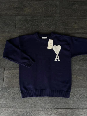 Mörkblå stickad tröja från Ami - Snygg mörkblå stickad tröja från Ami med vit hjärtlogga och bokstaven A på bröstet. Tröjan har rund hals, ribbade muddar och är långärmad. Perfekt för dig som gillar stilrena detaljer och vill ha något som sticker ut lite extra.