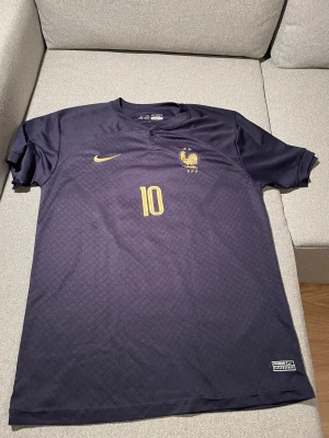 Frankrike Mbappé Nike fotbollströja XL - Säljer en officiell Frankrike fotbollströja från Nike med Mbappé och nummer 10 i guld. Tröjan är mörkblå med subtilt rutmönster, broderad tupp-logga och Nike swoosh. Tillverkad i lätt, ventilerande Dri-FIT polyester. Perfekt för dig som älskar landslagsfotboll.