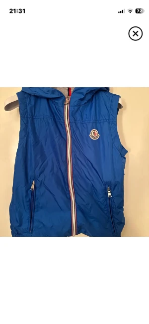 Blå Moncler dunväst med huva - Snygg blå dunväst från Moncler med grå huva och dragkedja framtill. Västen har två fickor med dragkedja och Moncler-logga på bröstet. Insidan är grå och materialet känns lätt och sportigt. Perfekt för lager-på-lager och streetstyle.