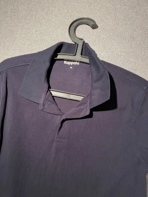 Mörkblå pikétröja från Kappahl - Snygg mörkblå pikétröja från Kappahl i storlek M. Klassisk krage och korta ärmar, tillverkad i mjuk bomull som är skön mot huden. Perfekt för dig som gillar enkel och stilren look. Passar dig som vill ha en basic piké med bra passform.