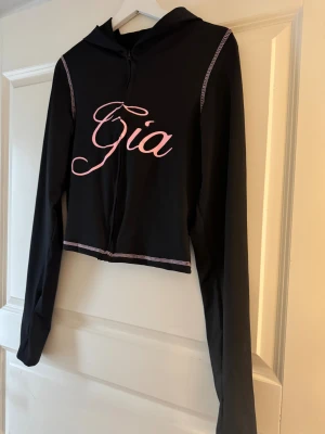 Svart croppad hoodie med rosa tryck - Svart croppad hoodie från Gia med rosa tryck över bröstet och kontrastsömmar i rosa. Tröjan har lång ärm, huva och dragkedja framtill. Perfekt för dig som gillar streetstyle och vill sticka ut med coola detaljer.