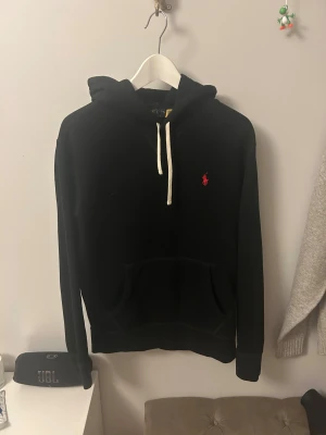 Svart hoodie från Polo Ralph Lauren - Svart hoodie från Polo Ralph Lauren med vit snörning och röd broderad logga på bröstet. Klassisk känguruficka framtill och mjukt material som känns skönt mot huden. Perfekt för dig som gillar stilren streetwear med premiumkänsla.