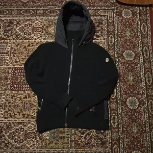 Svart dunväst med huva från Moncler - Snygg svart dunväst från Moncler med stickade sidor och ärmar, quiltad framsida och baksida samt stor vadderad huva. Västen har dragkedja framtill och Moncler-logga på ärmen. Perfekt för lager-på-lager och streetstyle.