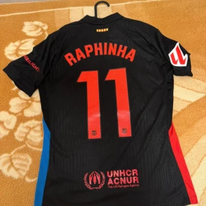 FC Barcelona Raphinha svart matchtröja - Säljer en svart FC Barcelona fotbollströja med Raphinha och nummer 11 på ryggen. Tröjan har röda och blå detaljer på sidorna, Nike-logga, Spotify och UNHCR-tryck samt klubbmärke framtill. Tillverkad i lätt och ventilerande material.