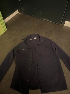 Mörkblå overshirt från Selected Homme - Stilren mörkblå overshirt från Selected Homme med klassisk krage, två bröstfickor med knappar och raka linjer. Jackan har tryckknappar framtill och är tillverkad i ett mjukt material som passar perfekt för lager-på-lager. Enkel och clean look.