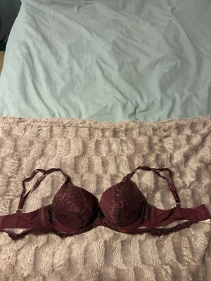 Vinröd spets bh med svarta detaljer - Säljer en vinröd spets bh med svarta spetsdetaljer från Hunkemöller. Den har vadderade kupor, justerbara axelband och en liten svart rosett framtill. Materialet är en blandning av spets och mesh som ger en lyxig känsla.