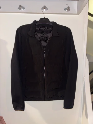 Svart Zara cardigan med dragkedja - Svart cardigan från Zara med enkel design och hel dragkedja framtill. Jackan har ståkrage, långa ärmar och quiltad struktur.